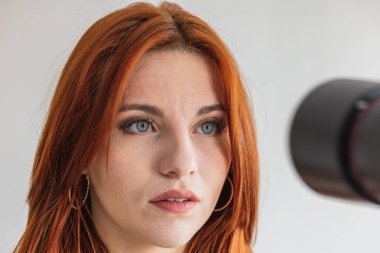 Kamera merceği yüzüne doğrultulmuş bir fotoğraf çekimi sırasında beyaz bir model. Kızıl saçlı, mavi gözlü, beyaz arka planda soyutlanmış genç bir kadın..