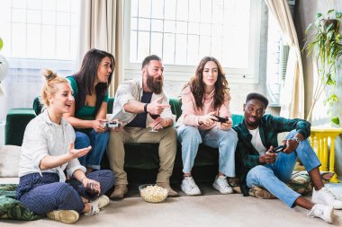 Çoklu etnik çeşitlilikte karışık arkadaşlar video oyunları oynayarak eğleniyorlar. Kahkahalar ve gülücükler elinde kontrollerle koltukta oturuyor.