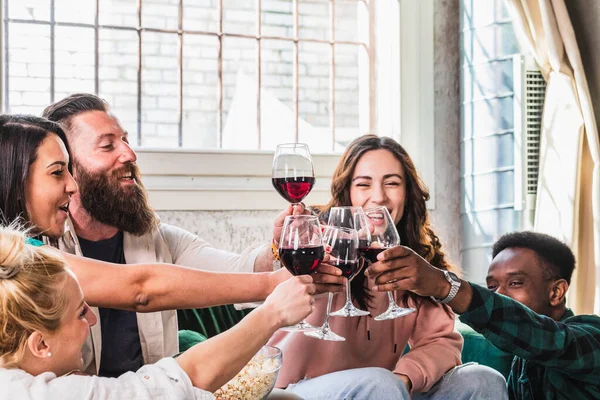 Cheers! People celebrate and raise glasses of red wine for toast. Focus on glasses. Group of happy man and woman cheering and having fun indoor in living room.