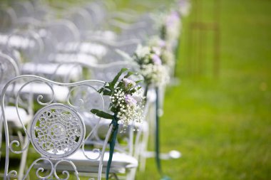 Açık havada düğün töreni için yeşil çimlerde duran çiçeklerle süslenmiş beyaz sandalyeler.