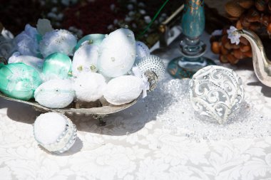 Süslü yumurta ve küreler gümüş tepsi ve cam vazo ile beyaz bir masa örtüsü ile kaplı bir masanın üzerinde yatmaktadır.