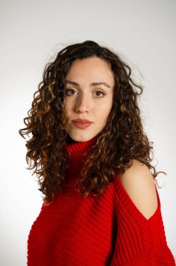 Uzun kıvırcık saçlı genç kadın kırmızı kazak içinde poz veriyor.