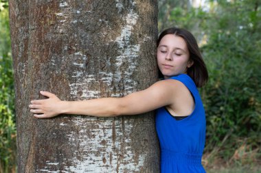 Genç kadın, gözleri kapalı büyük bir ağaca sarılarak doğanın kalbinde huzuru buluyor.