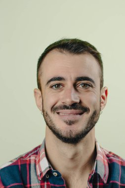 Sakallı ve ekose gömlekli genç adam yeşil bir arka planda gülümsüyor.