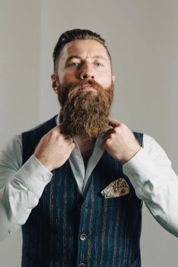 Uzun sakallı şık bir işadamı stüdyo ortamında dikilirken yeleğinin düğmelerini ilikliyor.