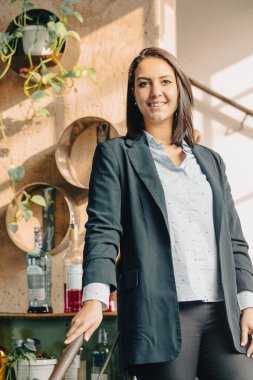 Ofisinde bitki ve şişelerle dikilirken gülümseyen bir iş kadınının portresi.