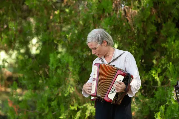 Kıdemli bayan müzisyen ormanda duruyor ve kırmızı akordeonda müzik çalıyor.