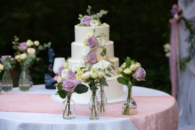 Üç katlı, güllerle süslenmiş, açık havada pembe bir masa örtüsü olan bir masada duran bir düğün pastası.