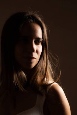 Uzun kahverengi saçlı, beyaz kolsuz tişörtlü dramatik ışıklandırma ve gölgelendirme ile vurgulanmış genç bir kadının stüdyo portresi.