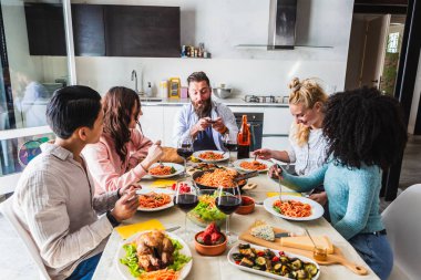 Bir grup çok ırklı arkadaş evde spagetti ve kırmızı şarap içerken, biri akıllı telefonuyla fotoğraf çekiyor.