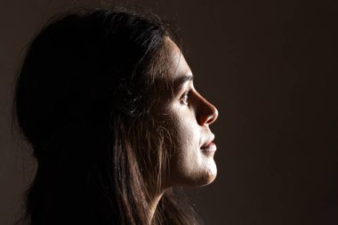 Yukarıya bakan genç bir kadının profili, yukarıdan gelen bir ışık kaynağıyla aydınlatılmış, karanlık bir arkaplanda.