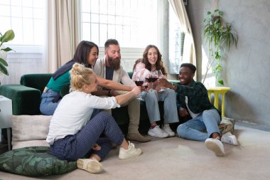 Bir grup neşeli çoklu etnik arkadaş kırmızı şarap içerken evde birlikte yerde ve kanepede oturuyorlar.