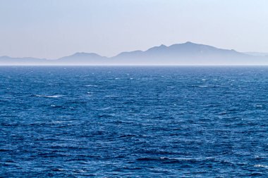 Açık bir günde engin mavi okyanus ufka doğru uzanırken arka planda puslu dağlar.