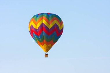 Parlak renkli sıcak hava balonu, huzurlu mavi gökyüzüne doğru zarif bir şekilde süzülür. Hava yolculuğunun resimli bir görüntüsünü sunar.