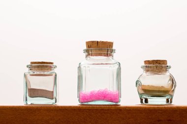 Kum ve pembe kristaller içeren üç cam kavanoz beyaz arka plana karşı ahşap bir rafta duruyor ve basit ama zarif bir hayat yaratıyor.