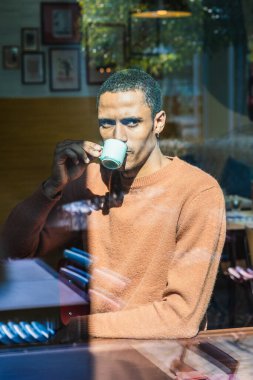Bir kafede kahve içen genç adam, pencereden görülüyor.