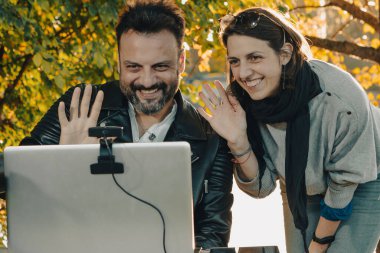 Bir adam ve bir kadın parkta dizüstü bilgisayardan kameraya el sallıyor.