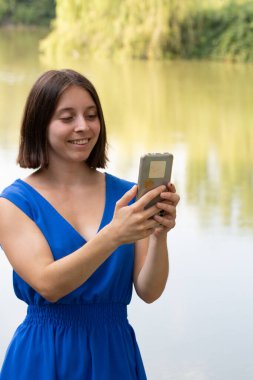 Göl kenarında akıllı telefon kullanan bir kadın, cep telefonu teknolojisinden ve doğadaki bağlantıdan zevk alıyor.