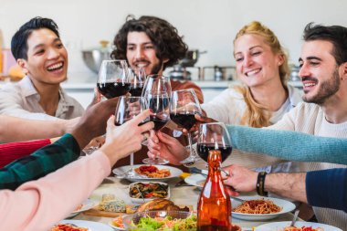 Neşeli çoklu etnik arkadaşlar evde İtalyan yemeğinin tadını çıkarırken kırmızı şarap kadehleriyle kadeh kaldırıyorlar.