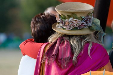 İki kadın bir açık hava etkinliğinde hassas bir anı paylaşıyor, biri kendine özgü bir çiçek şapkası takıyor.
