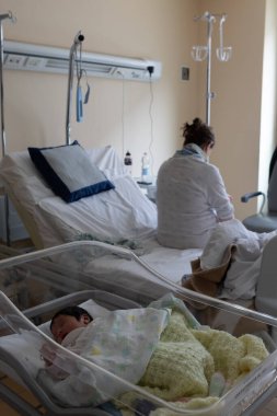 Yeni doğmuş bebek hastane beşiğinde huzur içinde uyurken annesi de yakınlardaki yatakta oturuyor.