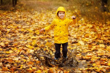 Sonbahar parkında su birikintisine atlayan sarı yağmurluk ve lastik çizmeli mutlu çocuk. Her havada çocuklar için açık hava eğlencesi kavramı. Çocuklar için yağmur geçirmez giysi