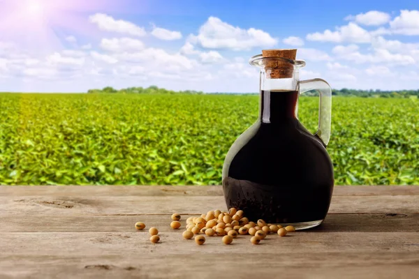 soy sauce in glass bottle with dry soybeans on wooden table and green agriculture field on the background