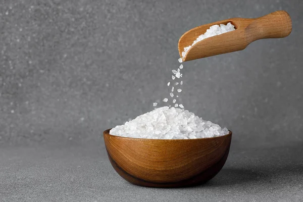 sea salt crystals falling from wooden scoop in bowl on grey background