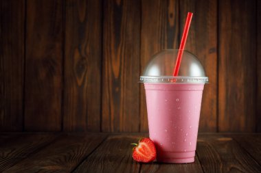 Tek kullanımlık plastik bardakta çilekli milkshake ve ahşap arka planda saman.