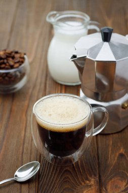 Cam fincanda espresso, gayzer kahve makinesi ve tahta masada bir sürahi krema.