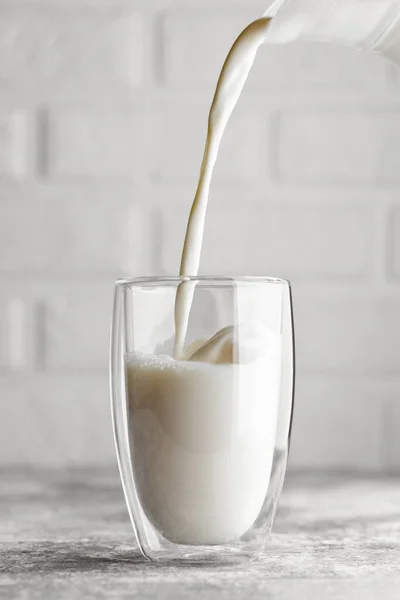 milk pouring in glass from jug on grey table with white brick wall as background
