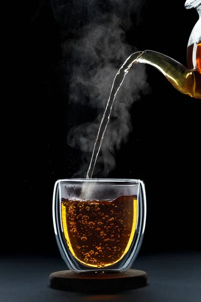 stock image hot tea pouring from teapot into double wall glass cup on black background