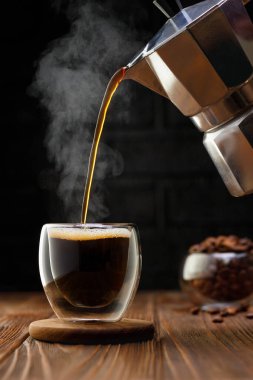 Sıcak espresso, gayzer kahve makinesinden siyah arka planlı, ahşap masada cam fincana dökülüyor.