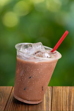 Eğik, tek kullanımlık çikolatalı milkshake. Arka planda yeşil bahçe ve ahşap masa üzerinde buz.