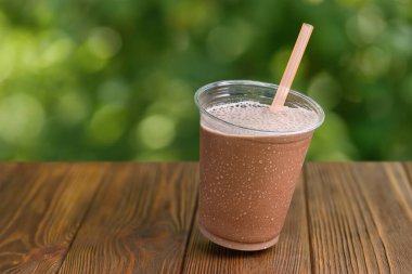 Arkadaki yeşil bahçeli ahşap masaya yatırılmış plastik bardak çikolatalı milkshake.