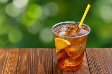 Tek kullanımlık plastik bardak, buzlu çay ve limonlu ahşap masa ve arka planda yeşil bahçe.