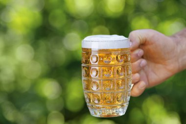 Yakın plan erkek eli köpüklü hafif bira bardağını tutuyor ve yeşil doğal arka planda tost yapıyor.