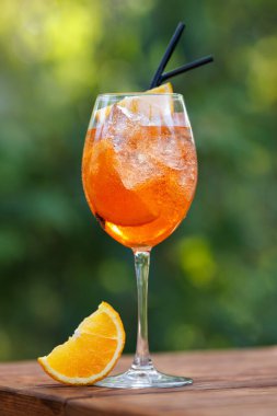 Siyah pipetli soğuk aperol spreyli kokteyl ve yeşil doğal arka planlı ahşap masada bir dilim portakal.