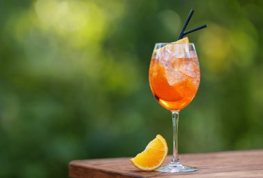 Siyah pipetli soğuk aperol spreyli kokteyl ve yeşil doğal arka planlı ahşap masada bir dilim portakal.