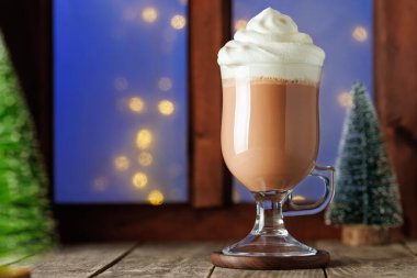 Sıcak çikolata ya da cam bardakta krem şantili kakao. Ahşap masada, arka planda pencere var.