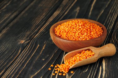 Kırmızı mercimek, kase dolusu tohumun yanındaki ahşap kepçeden dökülüyor ve koyu dokulu bir masada kırsal ve sağlıklı bir yemek sahnesi yaratıyor.