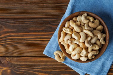 Cashew nuts are presented in bowl placed on blue napkin on rustic wooden table, creating warm and inviting scene perfect for food and lifestyle imagery. Top view