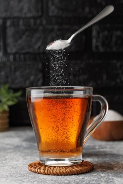 Çay kaşığından dökülen beyaz şeker hasır bardak altlığına konmuş sıcak çay bardağına dönüşüyor. Arka planda siyah tuğla duvara karşı çanak ve küçük bitki var.
