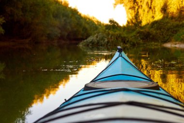 Tuna nehrinin kıyısında batan güneşin ışınlarıyla aydınlatılan ağaçlardan ve yabani üzümlerden oluşan yoğun yeşil çalılıklara karşı mavi kano (yay, burun).