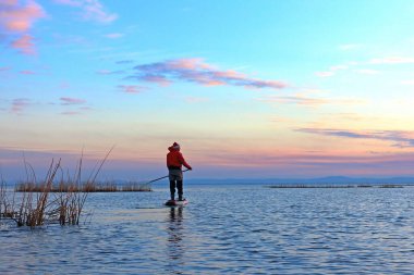 Man paddle (floating) on Stand up paddle board (SUP ) in Santa Claus costume in calm of winter lake at sunset clipart