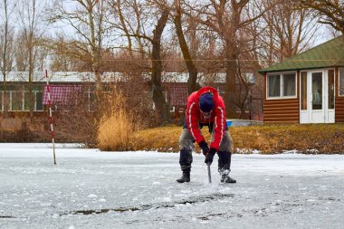 Kırmızı ceketli bir adam donmuş nehirdeki buzu baltayla deliyor.