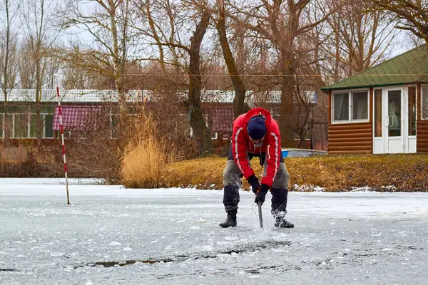 Kırmızı ceketli bir adam donmuş nehirdeki buzu baltayla deliyor.