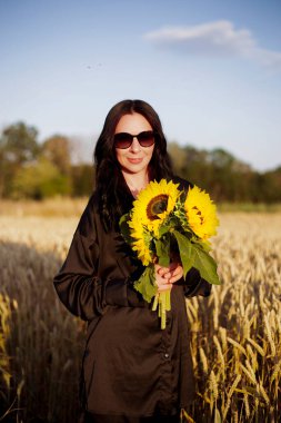 Elinde bir buket ayçiçeği olan siyah elbiseli bir kadın gün batımında dikenli bir tarlada duruyor. Yüksek kalite fotoğraf