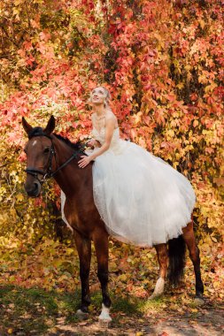 Beyaz uzun elbiseli güzel gelin sonbahar ormanında bir atın yanında duruyor. Doğada düğün. Yüksek kalite fotoğraf
