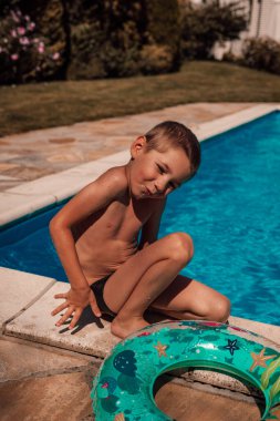 Küçük bir çocuk havuzun yanında oturuyor. Yaz sıcağında güneş yanığı, öğlen. Yüksek kalite fotoğraf